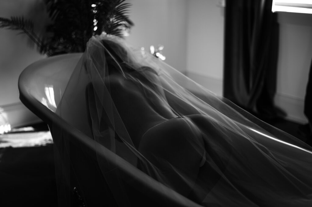 Black and white photo of a woman lying in a bathtub, draped in a sheer bridal veil, creating an intimate and artistic atmosphere with soft lighting.